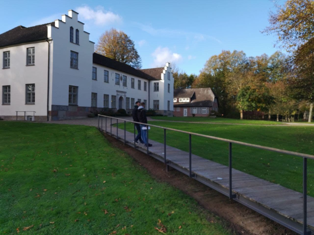 Ein weißes Herrenhaus, davor ein Holzsteg, auf dem zwei Menschen stehen.